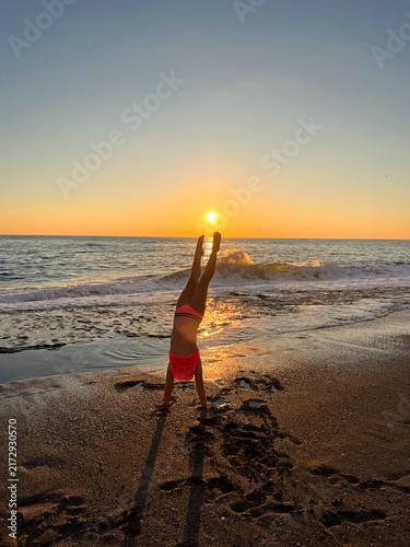 Active little girl doing a handstand on a sandy sea beach against a beautiful golden sunset sky with breaking waves