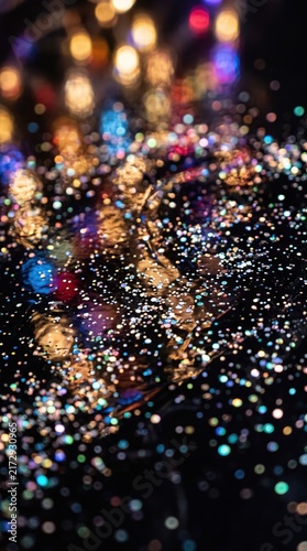 Glittering confetti scattered on a dark surface for a new year celebration. Festive lights create a glowing bokeh effect in the background.