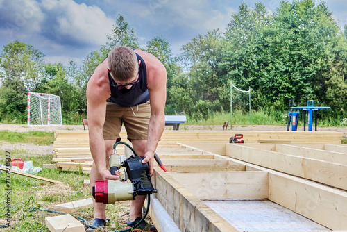 Quadro su tela Nail gun fastens wooden boards during outdoor floor frame construction, worker holds pneumatic tool near timber beam
