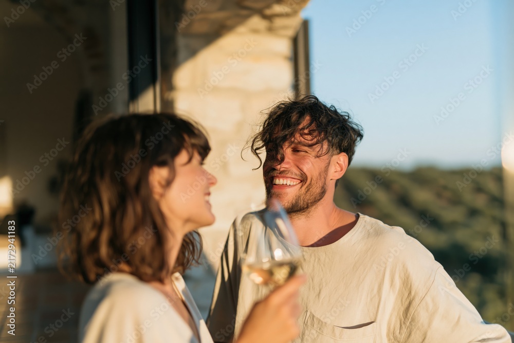 Naklejka premium Smiling couple drinking white wine on a sunny terrace overlooking hills for romantic lifestyle banner and travel background