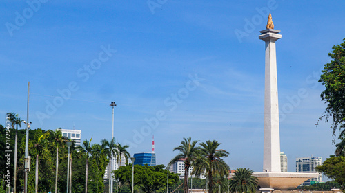 Fotografie The iconic National Monument (Monas) in Jakarta, Indonesia, is surrounded by a spacious plaza, green landscapes, palm trees, and modern city buildings beneath a bright blue sky
