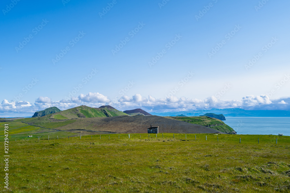Obraz premium A picturesque view of green grassy hills, a small building, and the ocean under a bright blue sky with fluffy clouds in Vestmannaeyjar, Iceland.