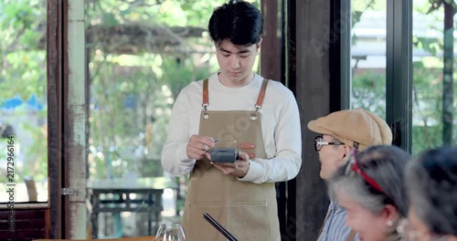 Young waiter using payment device beside senior asian diners in restaurant, preparing bill after reunion lunch, service moment with digital checkout, hospitality support, family style dining