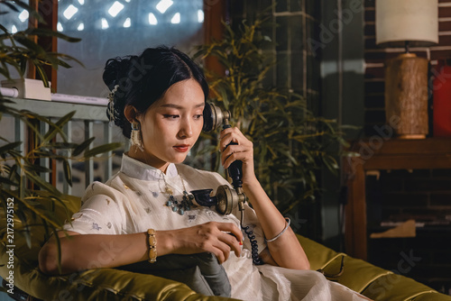 Photography Vintage styled Asian woman in qipao holding antique telephone receiver seated on