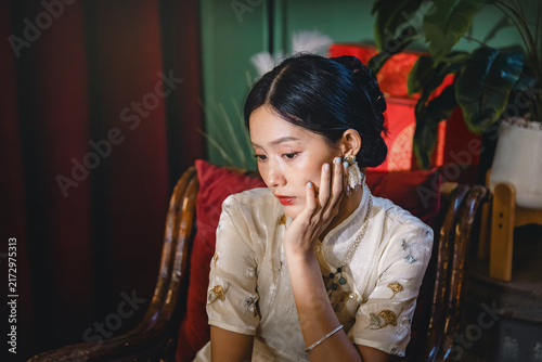 Photography Asian woman resting cheek on hand seated among red cushions in white embroidered