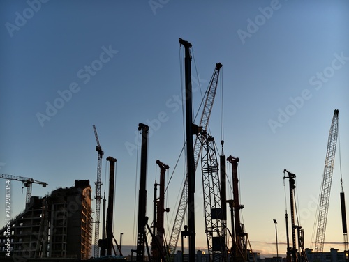 Construction cranes and piling rigs silhouetted against clear evening sky over urban building site, with tall machinery, unfinished structure, and sunset light creating industrial skyline