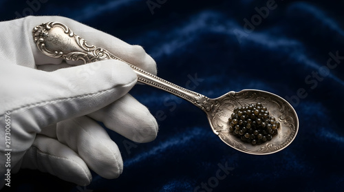 Hand wearing a white glove holding a vintage silver spoon filled with black caviar over a dark blue velvet background