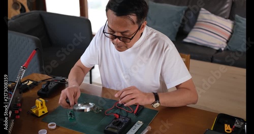 Senior Man soldering circuit board at home, electronics repair hobby engineering with hands on workbench assembly in cozy living room