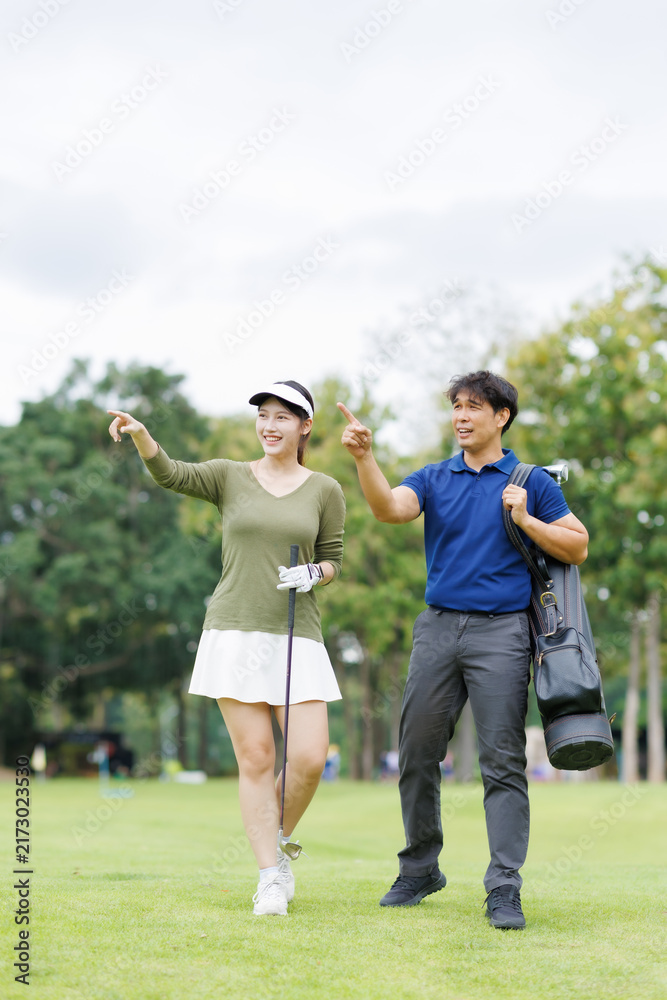 Obraz premium Two smiling Asian golf players walking on a green course, carrying clubs and a bag, pointing and chatting as they plan strategy and enjoy a sunny day outdoors