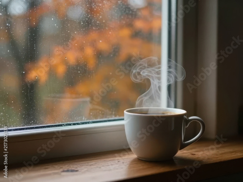 Ultra-realistic cozy morning scene with a steaming ceramic coffee cup beside a rainy autumn window, blurred orange foliage outside, warm interior light, cinematic lifestyle photography, soft atmospher