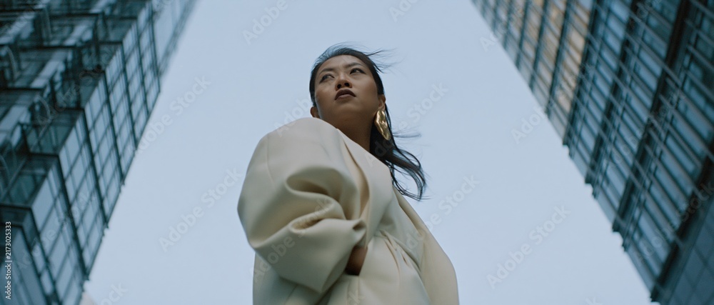 Naklejka premium Confident Asian woman looking up between towering city skyscrapers, symbolizing ambition and urban success