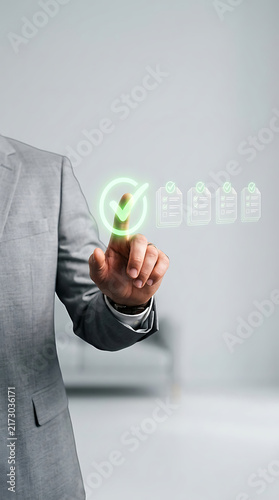Corporate worker in grey suit using holographic check mark symbol for document task approval and workflow management system interface within bright professional office workspace.