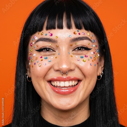 Creative face jewels on smiling woman, colorful rhinestone makeup portrait, vibrant orange background with festive glitter gems