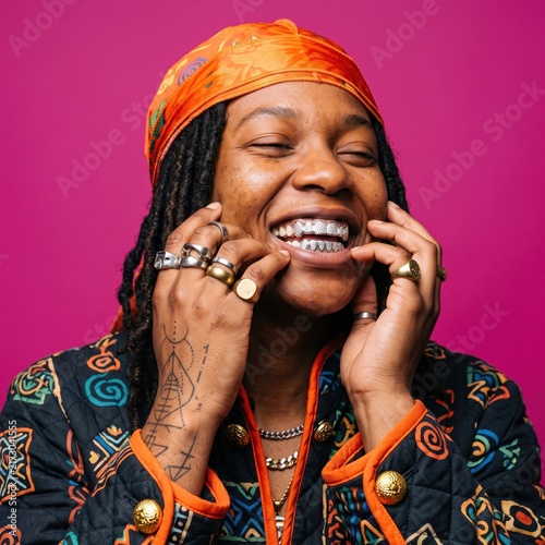 Stylish Black person with silver grillz and orange headscarf, happy individual with dreadlocks and gold rings, vibrant pink background portrait