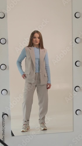 Vertical. Professional female model with long hair posing for a photo shoot in a studio, walking and looking at the camera while wearing a trendy business casual outfit reflected in a large mirror
