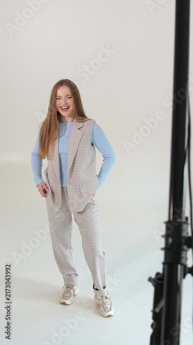 Vertical video. Playful young businesswoman in a striped suit and sneakers dancing and having fun in a professional photo studio with a white background and lighting equipment, isolated on white