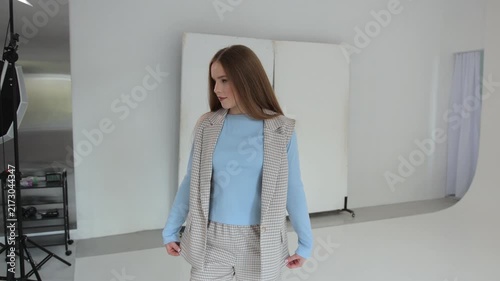 Beautiful young female model wearing a stylish plaid suit posing and dancing playfully in a professional photo studio with a white cyclorama and lighting equipment in the background