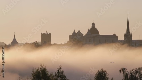 Dramatic foggy morning revealing the historic skyline of lutsk, ukraine, with the lubart's castle and saint peter and paul cathedral emerging from the dense, flowing mist at sunrise