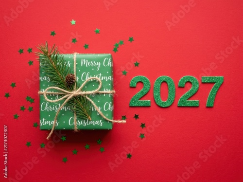 Christmas gift box wrapped in green paper with a pine branch and pinecone, next to 2027 glitter numbers on a red background with star confetti
