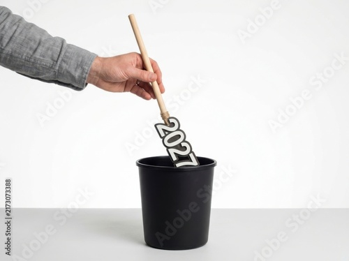 Hand placing a wooden sign with the year 2027 into a black trash bin, isolated on white background