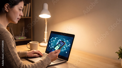 Person uses smartphone and laptop to check security features at a desk in an indoor setting during the evening