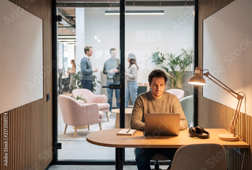 Man works on laptop in office while colleagues talk in background