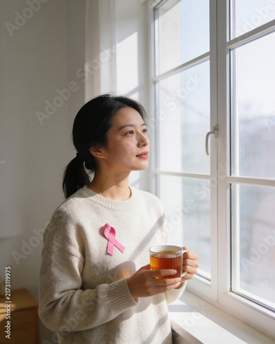 Asian woman wearing a sweater with a pink breast cancer awareness ribbon holding a warm cup of tea by a sunlit window. Hopeful, quiet reflection concept.