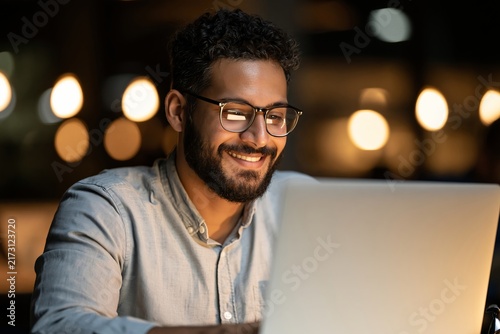 Smiling man with a beard and glasses using a laptop in a dimly lit room with soft glowing lights