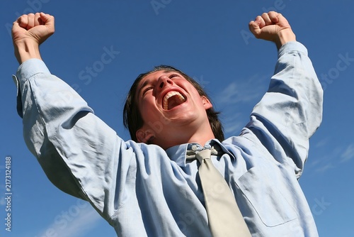Young businessman celebrating success with arms raised high against a clear blue summer sky background
