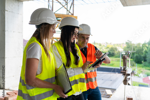 Inspectors and worker study technical details of project on tablet screen. Engineers and builder look at device with information about building