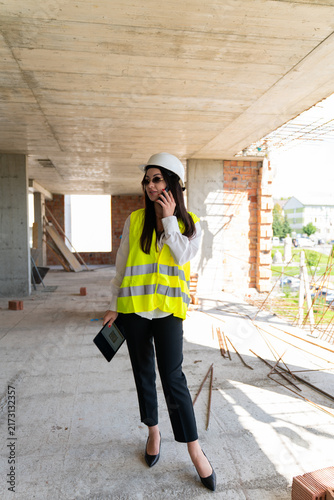 Lady in hard hat walks across raw floor talking on smartphone. Supervisor carries tablet and paces aisle confirming time slots while awaiting update