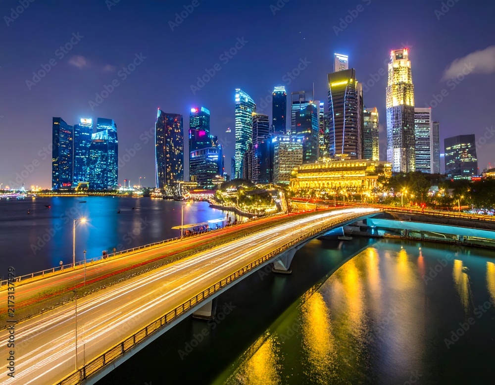 Fototapeta premium Nighttime cityscape with illuminated skyscrapers reflected in calm water; bridge with light trails