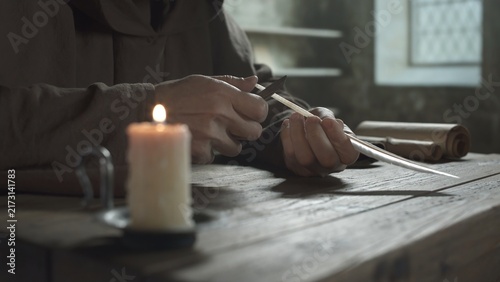 Konstfotografi Hands of a medieval monk cutting a feather quill pen with a knife at a wooden ta