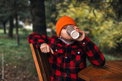 Young boy in a warm orange beanie drinking a hot beverage from a paper cup while sitting at a wooden table in a cozy cafe. Relaxing autumn lifestyle moment with a warm atmosphere