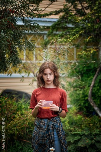 Teen girl standing in an outdoor garden, holding a plastic container filled with fresh orange sea buckthorn berries, looking directly at the viewer with a thoughtful expression