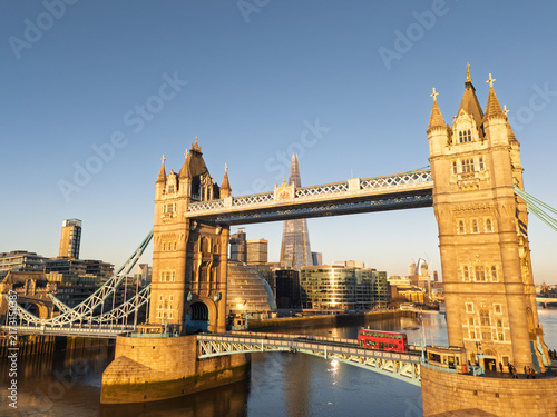 Aerial view of Tower Bridge in London at sunrise with a red double-decker bus crossing and The Shard in the background over the River Thames.