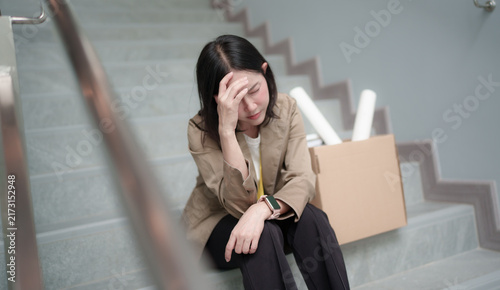 Obraz na płótnie Disappointed businesswoman sitting on office stairs beside a cardboard box, looking frustrated and uncertain about career change, relocation, or workplace challenges