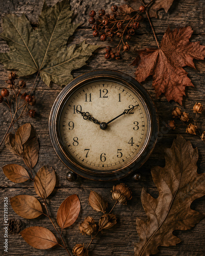 Vintage alarm clock rests peacefully surrounded by dry autumn leaves on a dark rustic wooden surface