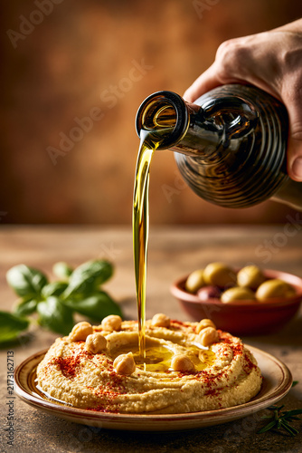 A dark glass bottle pouring a stream of golden-green extra virgin olive oil over a plate of homemade hummus garnished with chickpeas and paprika.