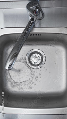 Hand Turning On Faucet to Fill Kitchen Sink. Top view of a person opening a chrome tap as running water begins filling a stainless steel sink.