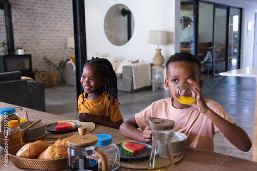 African American children sitting in open-plan home at wooden dining table drinking orange juice