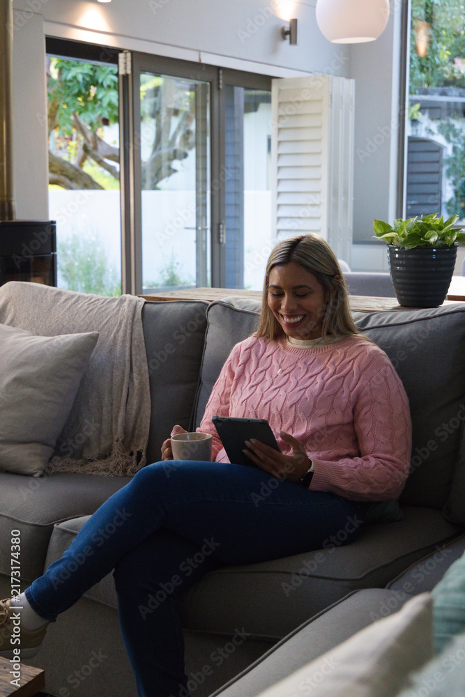 Naklejka premium Mid-adult African American woman sitting on grey sofa in living room holding tablet and mug