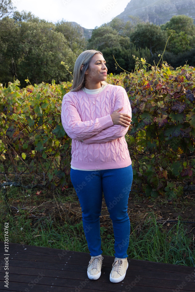 Fototapeta premium African American woman standing on wooden boardwalk in vineyard, wearing pink sweater and jeans