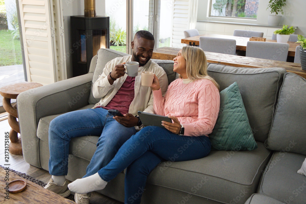 Naklejka premium Diverse couple sitting on gray sofa in living room holding mugs, tablet, smartphone, smiling