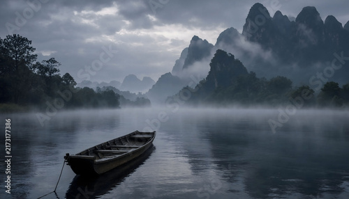 Silhouette of traditional wooden boat on calm misty lake with misty mountain backdrop
