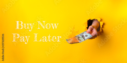 A hand extends a dollar bill through a torn yellow paper background, accompanied by the text 'Buy Now Pay Later', symbolizing modern consumer finance options.