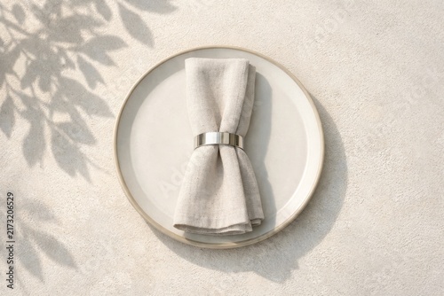 Minimal beige linen napkin folded with metal ring on round ceramic plate and soft leaf shadows for neutral table setting background and food styling flatlay design
