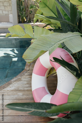 Pink Striped Pool Float by Tropical Poolside. Pink and white striped inflatable swim ring resting on a rustic wooden deck beside a turquoise swimming pool, surrounded by lush tropical banana leaves. 