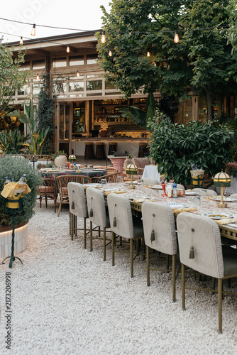 Italian Garden Restaurant with String Lights at Dusk. Dining area of a Mediterranean restaurant in the evening: elegant table setting with yellow napkins and patterned plates, vintage table lamps, bar