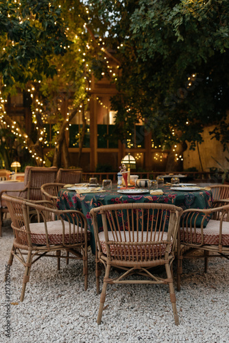 Romantic and Elegant Garden Dinner Table Under Fairy Lights at Dusk. Round dining table with dark green floral tablecloth and vintage rattan armchairs set for dinner in a garden restaurant courtyard. 